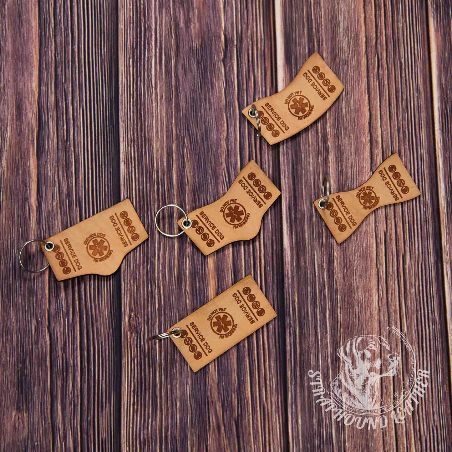 photo showing 5 service dog shaped and laser engraved keychains in natural leather with silver split rings depicting miniature replicas of service dog vests on a wood grain background